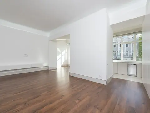 A clean, empty apartment room with white walls, wood-look flooring and large windows